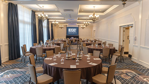 Medium-sized meeting room with blue curtains hung by large windows, rows of long tables, and a presentation screen situated at the front of the room.