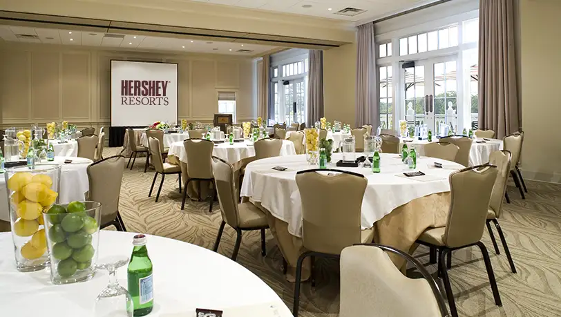Large meeting room room with nine tables neatly decorated with white tableclothes and topped with waters, fresh fruit, and Hershey bars on the tables.