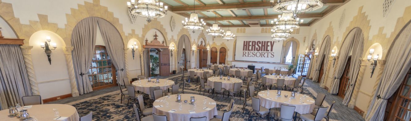 Room with large open windows and rows of long tables lined up with chairs, facing the front of the room.